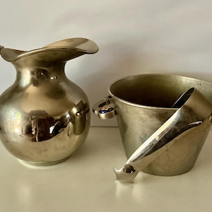 May include: A polished silver-tone metal pitcher with a curved handle and a matching ice bucket featuring side handles with blue and gold circular accents, accompanied by a silver-tone metal ice scoop resting inside the bucket.