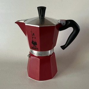 May include: A red aluminum Moka pot coffee maker with a black handle and lid knob. The body features the Bialetti logo and the text Moka Color printed on the silver-toned middle band. The octagonal design includes a spout and a hinged lid.