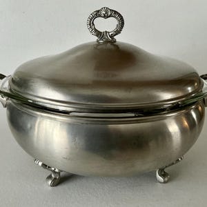 May include: A silver-colored serving dish with a lid and handles. The dish has a rounded shape and sits on three small feet. The lid has a decorative handle. The dish appears to be made of metal and glass.