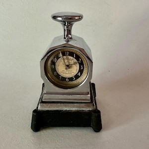 May include: A chrome-plated Orson Jr. desk clock featuring a plunger-style top handle, a circular face with black numerals on a cream background, and a dark, heavy rectangular base. The clock face displays the text Orson Jr. and patent numbers.