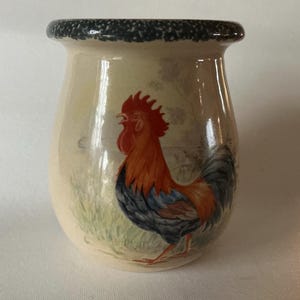 May include: A ceramic jar with a rooster design. The jar has a cream-colored base with a detailed illustration of a rooster in shades of red, orange, and blue. The rim is dark blue with a speckled pattern. The jar is likely used for kitchen storage.