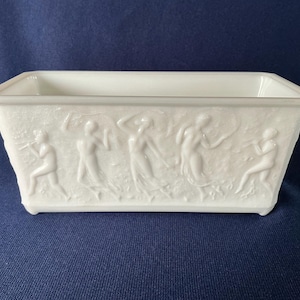 May include: A rectangular, white ceramic planter with a raised relief design. The relief depicts figures in classical poses. The planter has a smooth, glossy finish and sits on a blue surface.