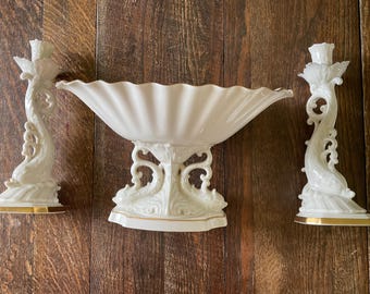 May include: A set of three white porcelain serving pieces with gold accents. The centerpiece is a large, scalloped bowl with a pedestal base. The two smaller pieces are candle holders shaped like dolphins.