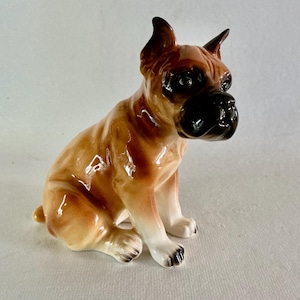 May include: A ceramic Boxer dog figurine in a seated position. The dog has a glossy finish with brown and tan coloring. The dog's snout and ears are black, and its paws are white with black markings.