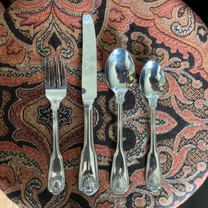 May include: A set of four silver-colored utensils: a fork, a knife, a spoon, and a smaller spoon. The utensils are arranged on a patterned surface with shades of brown, orange, and black. Each utensil has a decorative shell design on the handle.