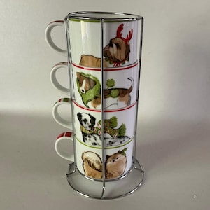 May include: A set of four stacked white ceramic mugs with dog illustrations, each adorned with festive accessories. The mugs are held in a silver metal rack. The mugs have red and green accents.