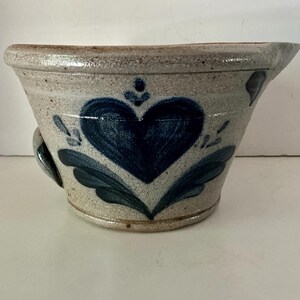 May include: A handmade stoneware bowl with a blue heart and leaf design. The bowl has a light beige background with a dark blue heart and leaf pattern. It has a small handle on the side.