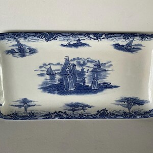 May include: A rectangular ceramic serving tray with rounded handles, featuring a blue and white transferware pattern. The design depicts a coastal scene with figures, sailboats, and a windmill, surrounded by a decorative floral border.