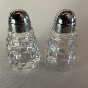 May include: A pair of clear glass salt and pepper shakers. Each shaker has a faceted, diamond-like pattern on the body and a silver-colored metal lid with small holes. The shakers are designed for tabletop use.