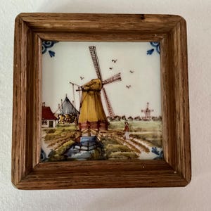 May include: A framed ceramic tile featuring a Dutch windmill scene. The artwork depicts a yellow windmill, a small house with a red roof, and a person walking on a path. The frame is made of wood, and the corners of the tile have blue floral accents.