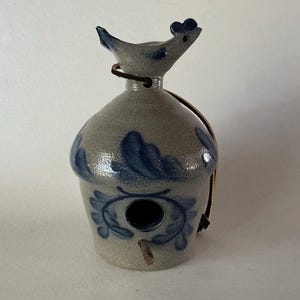 May include: A gray ceramic birdhouse with a glossy finish and blue painted floral patterns. The top features a sculpted bird figure, a circular entrance hole, a small wooden perch, and a brown leather hanging strap attached to the neck.