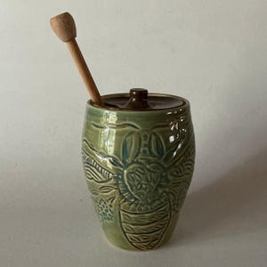 May include: A sage green ceramic honey pot featuring a relief-carved bee design on the exterior. The pot includes a dark brown circular lid with a small knob and a light-colored wooden honey dipper resting inside the opening of the lid.