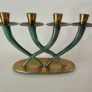 May include: A decorative candelabra with four candle holders. The base is an oval shape in gold, with four curved, teal supports. The candle holders are gold-colored and sit on small, round, teal platforms.