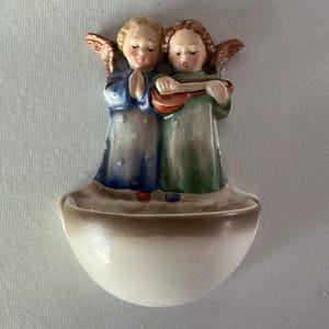 May include: A ceramic wall-mounted holy water font featuring two painted angels. One angel wears a blue robe with hands clasped in prayer, while the other wears a green robe and plays a small stringed instrument. Both have wings.