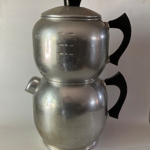 May include: A silver two-tiered coffee percolator with black plastic handles and a lid. The percolator has markings for measuring coffee.