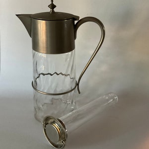 May include: A clear glass pitcher with a textured, wavy design, topped with a silver-colored metal lid and handle. A separate glass infuser with a silver-colored metal filter lies beside the pitcher. The pitcher is designed for serving beverages.