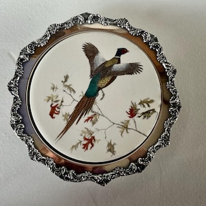 May include: A decorative coaster with a detailed illustration of a pheasant in flight, set against a white background with a branch and leaves. The coaster is framed by a silver-colored, ornate, scalloped edge.