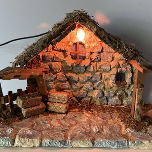 May include: A miniature stone-textured building with a thatched roof, featuring a small illuminated light bulb, a circular window, and a wooden structure on the side with stacked hay bales. The base is paved with stone-like blocks and scattered moss.