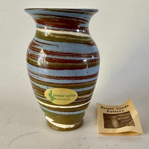 May include: A blue, brown, and white striped ceramic vase with a label that reads "Desert Sands Handmade".