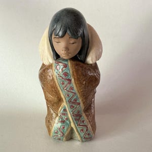 May include: A ceramic figurine of a person with dark hair and closed eyes, wearing a brown coat with a light-colored fur-lined hood. The coat features a central vertical panel decorated with a teal and red geometric pattern on a cream background.