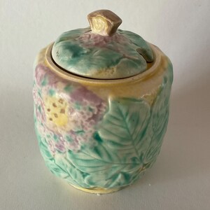 May include: A vintage ceramic sugar bowl with a lid. The bowl is decorated with a floral design in shades of green, yellow, and pink.