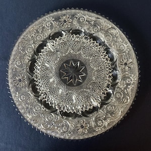 May include: A clear, decorative glass plate with intricate floral and starburst patterns. The plate features a scalloped edge and detailed engravings, creating a vintage aesthetic. The plate's design includes a central star motif.