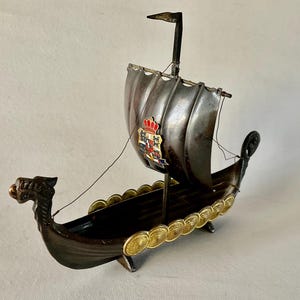 May include: A small, decorative metal ship with a dragon head prow, a sail with a crest, and a gold coin trim around the hull.