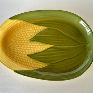 May include: Oval-shaped ceramic serving dish with a corn on the cob design. The dish features a yellow corn cob with green leaves. The dish is approximately 10 inches long and 6 inches wide. The dish has a glossy finish.