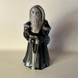 May include: A ceramic figurine of a robed figure with a long white beard, holding a brown deer. The figure wears a dark blue robe with white trim and a hood. The figurine is approximately 6 inches tall.