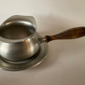 May include: A silver gravy boat with a wooden handle and a matching saucer. The boat has a rounded shape and a wide, shallow bowl.