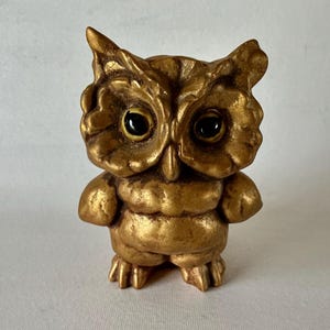 May include: A golden owl figurine with large, dark eyes. The owl has detailed feather textures and a slightly rounded body. The figurine is standing upright, showcasing its small feet and a textured surface. The owl is a decorative item.