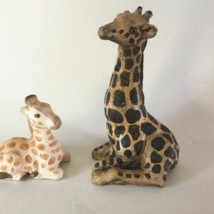 May include: Two ceramic giraffe figurines, one larger than the other. The larger giraffe is brown and black with a spotted pattern. The smaller giraffe is white and brown with a spotted pattern.