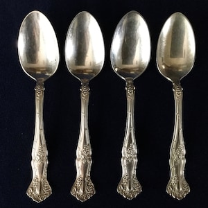 May include: Four silver teaspoons with ornate handles featuring a grapevine design. The spoons have a polished finish and are in good condition.