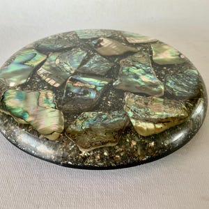 May include: Round, black coaster with iridescent green, blue, and white abalone shell pieces embedded in the surface.