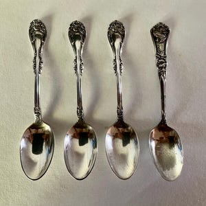 May include: Four antique silver spoons with ornate floral designs on the handles. The spoons have a reflective, polished surface, and are arranged on a light-colored background. The spoons are likely for serving or eating.
