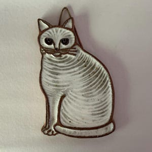 May include: Ceramic cat figurine in a seated position. The cat is white with gray stripes and brown trim. The figurine has a loop at the top for hanging. The cat's eyes are black, and it has a small brown nose and whiskers.