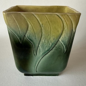 May include: A square ceramic vase with a leaf design. The vase transitions from a dark green at the base to a yellow-green at the top. The leaf pattern is raised and adds texture to the vase.