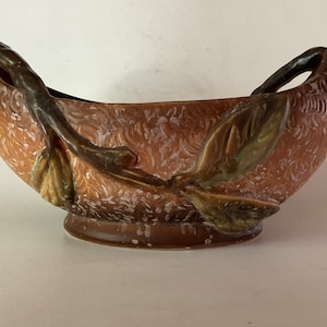May include: A brown ceramic bowl with two handles shaped like branches. The bowl has a textured surface and is decorated with green leaves.