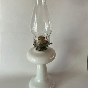 May include: An oil lamp featuring a smooth, opaque white glass base and a matching white glass font. The lamp includes a clear, bulbous glass chimney that sits atop a metallic burner assembly with a small adjustment knob on the side.