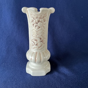 May include: A small, off-white ceramic vase with a fluted top and a pedestal base. The vase features embossed floral designs in a darker shade of the same color. The vase is approximately 6 inches tall.