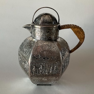 May include: A silver teapot with an ornate design and a brown wooden handle. The teapot has a round body with a lid and a spout. The design on the teapot is intricate and includes floral motifs and figures.