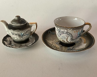 May include: A gray and white tea set with a dragon design. The teapot has a lid and a small spout. The teacup and saucer have a matching design.