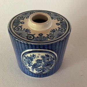 May include: A blue and white ceramic container with a floral design. The cylindrical body has vertical ridges and a decorative oval with a bird and floral motif. The top has a circular design and a central opening.