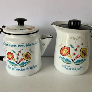 May include: Two white enamelware coffee pots with black handles and lids. The pots are decorated with colorful floral designs and text in blue. One pot has a spout and the other has a pitcher shape.