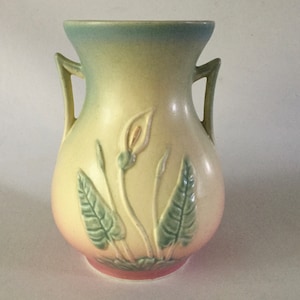 May include: A vintage glazed ceramic vase with two handles. The vase is decorated with a floral design of white calla lilies and green leaves. The vase has a light blue, yellow, and pink gradient.