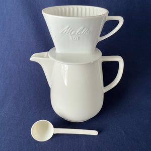 May include: A white ceramic pour-over coffee maker set, including a filter holder, a carafe, and a scoop. The filter holder is marked with the brand name "Melitta 101". The set is designed for brewing coffee.