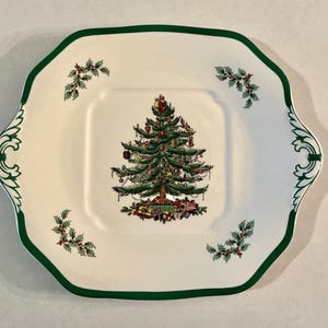 May include: A white, square-shaped Christmas serving platter with a green scalloped edge. The center features a decorated Christmas tree with presents. Holly sprigs are placed in each corner and on the handles.
