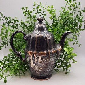 May include: Antique silver-plated teapot with a dark, aged finish. The teapot features a curved handle, a spout, and a decorative lid. The design includes vertical ridges and a band of copper-colored metal around the middle. The teapot is a vintage item.
