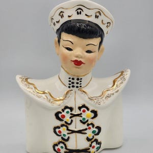 May include: A vintage ceramic figurine of a person in a white uniform with gold trim. The figurine has black hair, red lips, and a white hat. The uniform has decorative floral accents in red, green, and yellow.