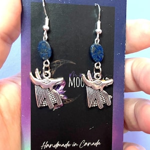 May include: Silver earrings with a blue lapis lazuli stone and a silver Anubis head charm. The earrings are handmade in Canada.